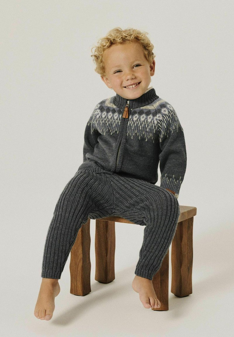 Bambino sorridente con capelli biondi ricci, che indossa un maglione e pantaloni in maglia grigio scuro, seduto a piedi nudi su uno sgabello di legno.