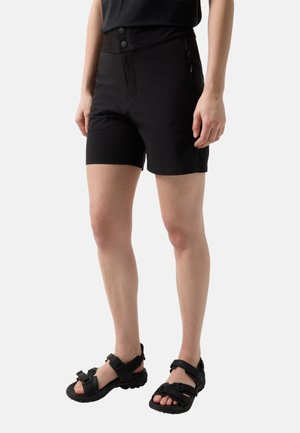 Osoba ubrana w czarne szorty z wysokim stanem zapinane na dwa guziki oraz czarne sandały na otwartym palcu z regulowanymi paskami, stojąca na białym tle.