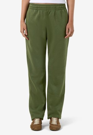 Personne portant un pantalon de survêtement ample vert olive et des baskets marron, debout devant un fond blanc uni.