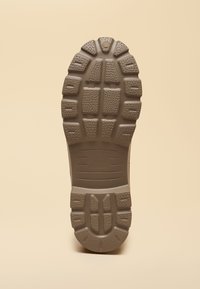 Sole of a brown boot with deep treads and textured grip pattern on a neutral beige background.