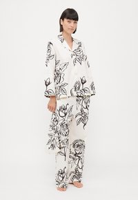 Set de pijama alb cu imprimeu floral negru, cu mâneci lungi și pantaloni largi. Material neted, croială ușor lejeră și design cu nasturi.
