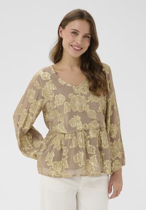 Mosolygó, hosszú hajú nő bézs blúzt visel arany virágmintákkal és hosszú ujjakkal, fehér nadrággal párosítva, kezét csípőjén tartva áll.