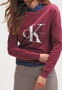 Sudadera burdeos con cuello redondo y logo "CK" en blanco. Tejido suave, puños acanalados, llevada sobre una camisa de mezclilla. Textura lisa y corte casual.