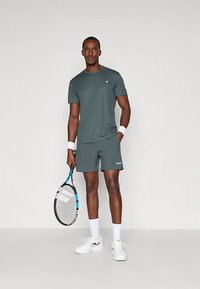 T-shirt atlético verde-escuro de mangas curtas, combinado com calções correspondentes. Jogador segura uma raquete de ténis, usando sapatilhas brancas e pulsos.