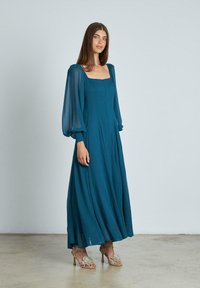 Teal Maxikleid mit quadratischem Ausschnitt, langen, durchsichtigen Puffärmeln und fließender Silhouette. Kombiniert mit dekorierten Sandalen. Glatte Textur.