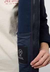 Marineblauwe jas met een fleece binnenvoering, voorzien van een rits en een label dat een 3-in-1 systeem beschrijft. De stof is glad met contrasterende texturen.
