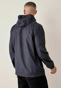 Mężczyzna w lekkiej czarnej kurtce z kapturem, stojący tyłem na jednolitym jasnym tle.