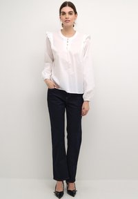 Blusa di cotone bianca con spalle arricciate e dettaglio a tre bottoni, abbinata a jeans blu scuro a gamba dritta e décolleté nere a punta.