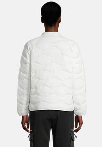 Veste matelassée blanche avec un col rond et des manches longues, présentant un motif en vagues. Portée par-dessus un pantalon noir avec des poches cargo.
