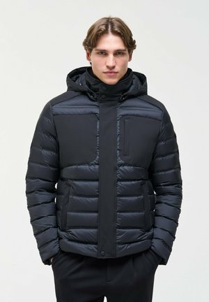 Un homme est de face, vêtu d’une veste matelassée foncée à capuche et d’un pantalon noir, sur un fond clair et uni.