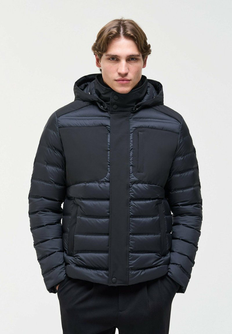 Un homme est de face, vêtu d’une veste matelassée foncée à capuche et d’un pantalon noir, sur un fond clair et uni.