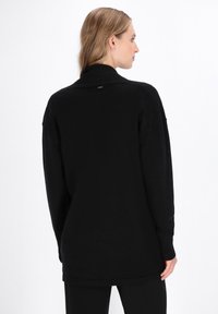 Schwarzer Pullover mit langen Ärmeln und Kragen, weicher Textur und lockerem Schnitt. Besitzt dezente Markenkennzeichnung auf der Rückseite und gerippte Bündchen.