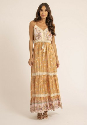 Femme debout portant une robe maxi fleurie sans manches avec des détails en crochet et des pompons, associée à des sandales à lanières.