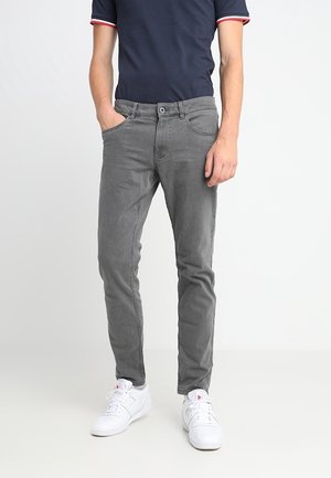 Homme portant un jean slim gris, un polo bleu marine et des baskets blanches, debout avec une main dans la poche.