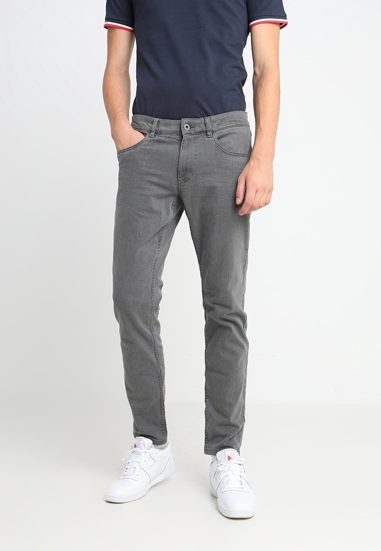 Homme portant un jean slim gris, un polo bleu marine et des baskets blanches, debout avec une main dans la poche.