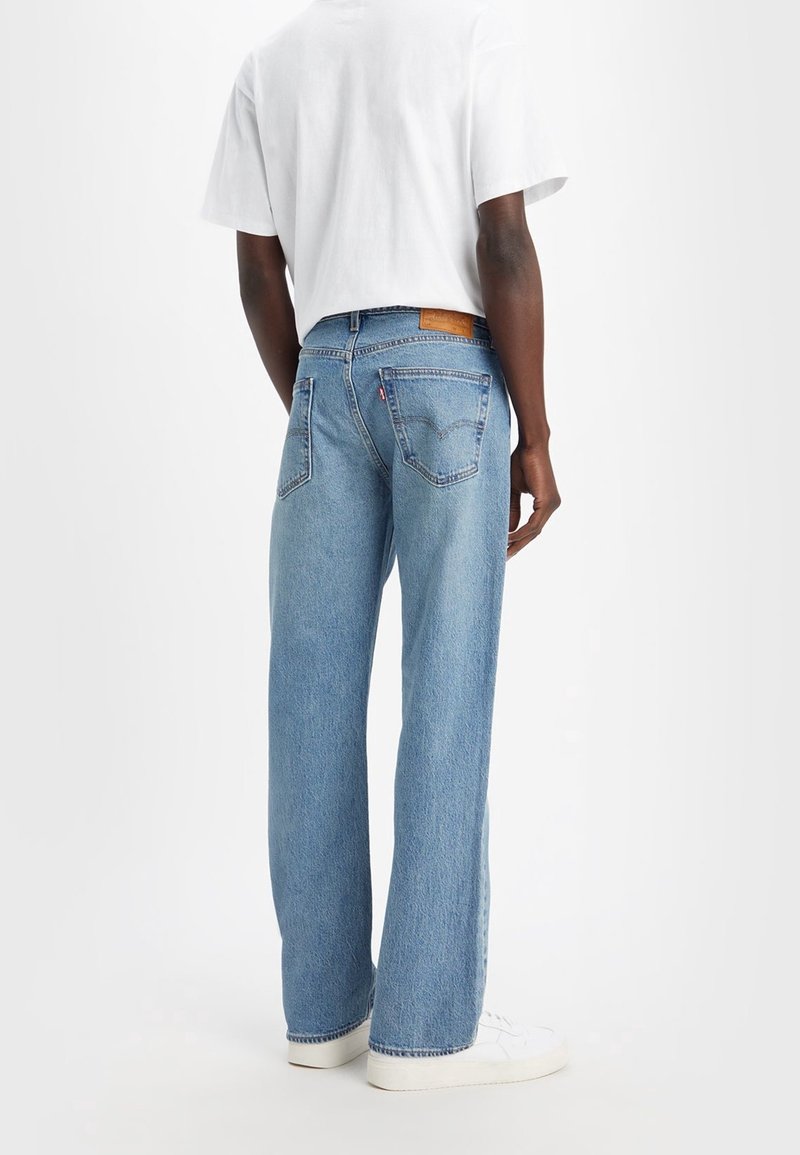 Jeans de mezclilla azul claro con corte recto, costuras visibles y un parche de cuero marrón en la cintura trasera. Combinados con una camisa blanca.