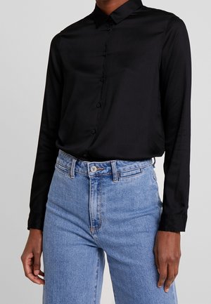 Osoba ubrana w czarną koszulę na długi rękaw zapinaną na guziki, wpuszczoną w jasnoniebieskie, wysokiej talii dżinsy z przodem zapinanym na guziki i szlufkami na pasek.