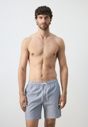 Oberkörperfreier Mann mit kurzen lockigen Haaren, der blaue und weiße, vertikal gestreifte Badeshorts mit Kordelzug trägt, vor einem schlichten hellen Hintergrund stehend.