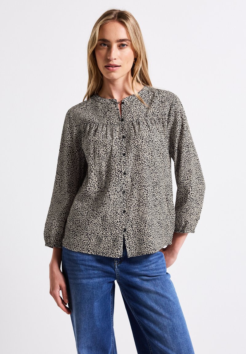 Beige Bluse mit schwarzem Tiermuster, mit Knopfleiste, gerafftem Ausschnitt und langen Ärmeln, kombiniert mit blauen Jeans.