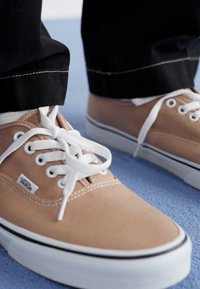 Beiga canvas-sneakers med vita snören, svarta sömmar och ett svart streck längs den vita gummisulans kant, som bärs med svarta byxor.