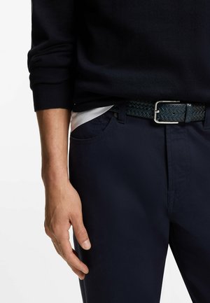 Marineblaue Hose mit einem strukturierten, gewebten schwarzen Gürtel. Der Pullover ist marineblau, die Bündchen sind sichtbar. Die Hand ruht auf der Hose und zeigt die Details des Kleidungsstücks.