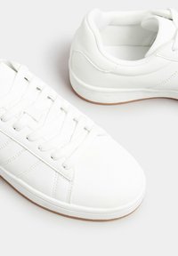 Vita idrottsskor i syntetläder med platta snören, en strukturerad sula och detaljer i söm på sidorna.