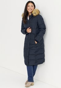 Donkerblauwe gewatteerde jas met een capuchon van imitatiewol, voorzien van een knoopsluiting aan de voorkant, een doorgestikt ontwerp en zijzakken; gecombineerd met een blauwe jeans en beige laarzen.