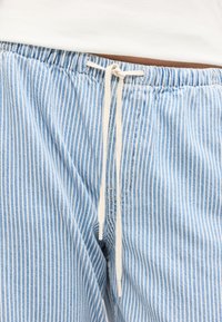 Μπλε και λευκό παντελόνι με κάθετες ρίγες και μπεζ κορδόνι στη μέση δεμένο σε φιόγκο, με λευκό τοπ που είναι μπαίνει μέσα στο παντελόνι.