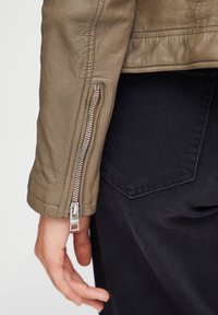 Light brown leather jacket featuring a zippered cuff, textured surface, and a close-fitting design paired with dark jeans and pocket details.