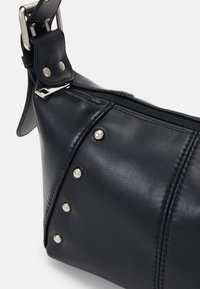 Sac bandoulière en cuir noir avec clous argentés, fermeture éclair et sangle ajustable avec boucle métallique sur fond blanc.