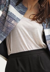 Veste à motifs bleus avec des rayures sur un haut beige clair, associée à un pantalon noir taille haute. Les tissus semblent doux et texturés.