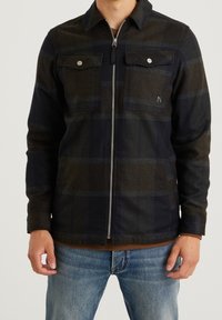 Muž v tmavé kostkované bundě na zip s kapsami na klopy na hrudi a modrých džínách, stojící před jednoduchým světlým pozadím.