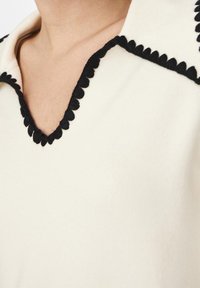 Weißes Oberteil mit tiefem V-Ausschnitt, eingefasst mit schwarzem gewelltem Detail, das eine glatte Stoffstruktur und ein maßgeschneidertes Design zeigt.