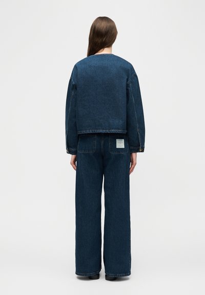 Άτομο που φοράει φαρδιά σκούρα μπλε τζιν μπουφάν και τζιν παντελόνι με φαρδιά πόδια, στέκεται με την πλάτη προς το μέρος του θεατή, πάνω σε απλό ανοιχτό φόντο.