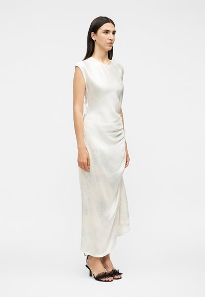 Γυναίκα με μακριά σκούρα μαλλιά που φοράει ένα λευκό σατέν φόρεμα χωρίς μανίκια και μαύρα ψηλοτάκουνα σανδάλια με φτερά, στέκεται μπροστά σε ένα απλό φόντο.