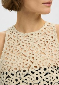 Beige hækletrøje med et detaljeret hulmønster med løkker. Stoffet går over til en ensfarvet sort kant forneden.