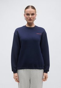 Tumesinine dressipluus pikkade varrukatega, soonikkoes ümara kaelusega ja alläärega. Rinnal punane logo. Pehme kangas ja mugav lõige.