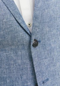 Jasnoniebieska marynarka z lnu o teksturowanej fakturze, zapinana na jeden czarny guzik, z klapami typu notch, zestawiona z białą koszulą z kołnierzykiem.