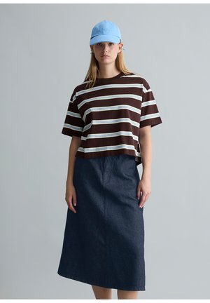 Giovane donna che indossa un berretto azzurro chiaro, una maglietta larga a righe marroni e bianche, e una gonna midi in denim blu scuro su uno sfondo grigio uniforme.