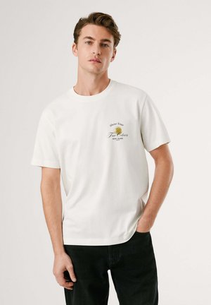 Jonge man in een wit T-shirt met korte mouwen met een klein geel blad en de tekst "Toon je ware kleuren", gecombineerd met zwarte broek, staand tegen een witte achtergrond.