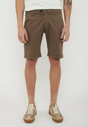 Pantaloncini casual marroni da uomo fino al ginocchio con orli arrotolati, indossati con sneaker beige e bianche con lacci e camicia bianca.