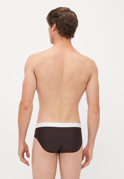 Braune Schwimmslips mit einem weißen elastischen Bund, enganliegendem Design und glatter Textur. Die Rückseite zeigt minimale Abdeckung und eine schlanke Silhouette.
