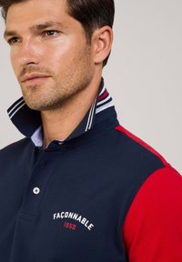Camisa polo azul marino con mangas rojas, con un cuello a rayas. Logo bordado "Faconnable 1950" en el pecho. Tejido texturizado.
