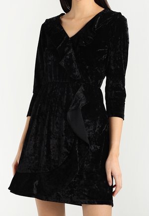 Zwarte fluwelen wikkeljurk met driekwart mouwen en ruches gedragen door een staand persoon tegen een effen witte achtergrond.