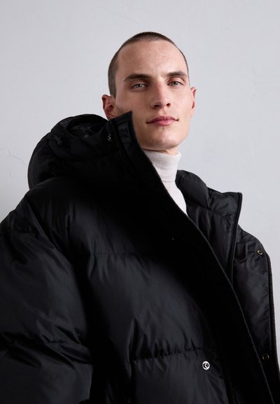 Jeune homme aux cheveux courts portant une doudoune noire et un col roulé clair sur fond gris uniforme.