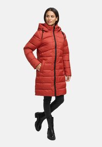 Kvinna iförd en lång röd vadderad kappa med huva, svarta tajta byxor och svarta kraftiga stövlar, står med en hand i fickan.