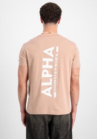 Krátky rukáv béžové tričko vyrobené z bavlny, so širokým bielym vertikálnym logom "ALPHA INDUSTRIES" na chrbte. Hladká textúra.