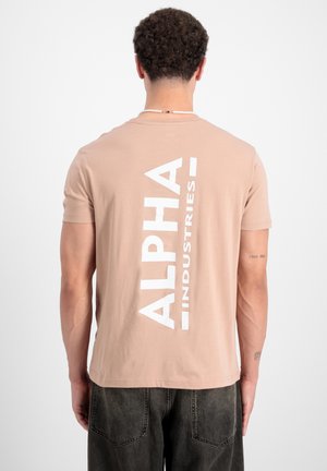 Bež majica kratkih rukava od pamuka s velikim bijelim vertikalnim logotipom "ALPHA INDUSTRIES" na leđima. Glatka tekstura.