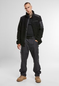 Giacca in pile nera con colletto alto e tasche con zip, abbinata a pantaloni cargo scuri e stivali beige, dal design utilitario.