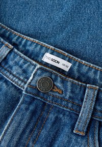 Traperice srednje plave boje, s bakrenim prošivanjem, crnim metalnim gumbom i bijelom etiketa na kojoj piše "TWO SOON, 28/32."